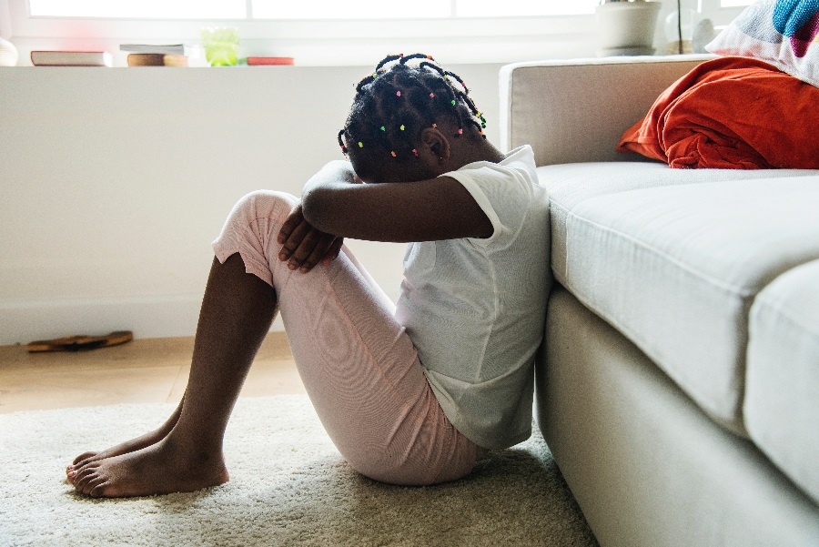 A young Black girl sitting in front of a couch with her head on her knees, showing sadness