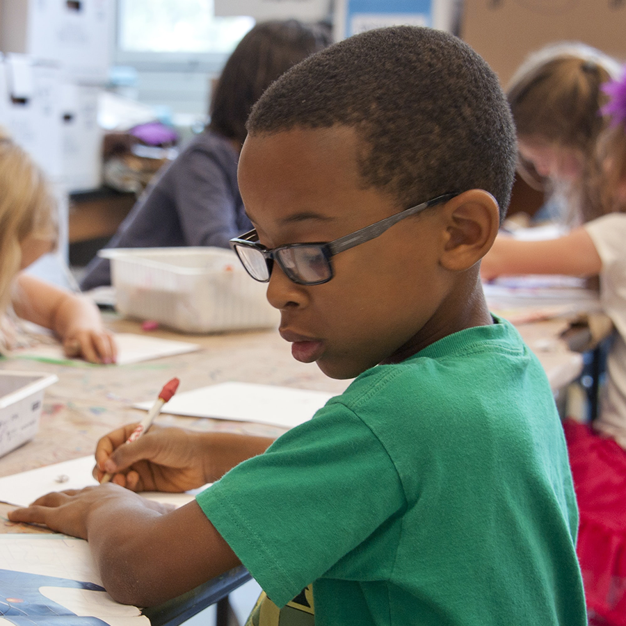 Kids working at a school table. The focus is on an African American boy in glasses