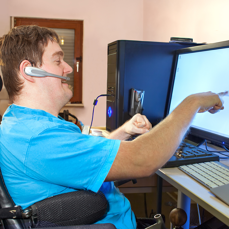 Man in a wheelchair wearing a blue shirt pointing at a computer screen.