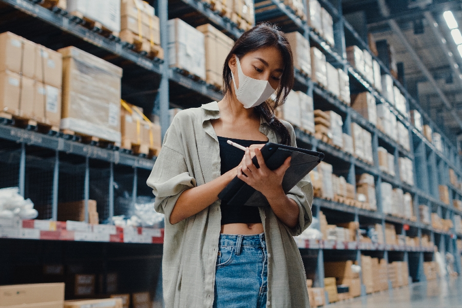 A young woman wearing a face mask in a warehouse while using a tablet