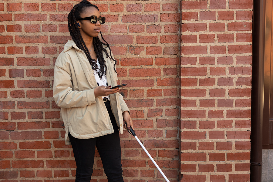African American woman who is blind standing in front of a brick wall, wearing sunglasses and holding a phone and cane