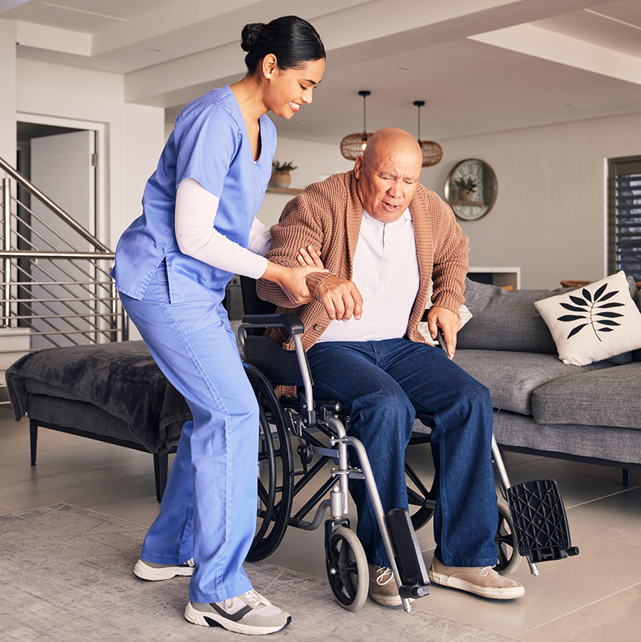 A nurse helps a man get out of a wheelchair in his living room