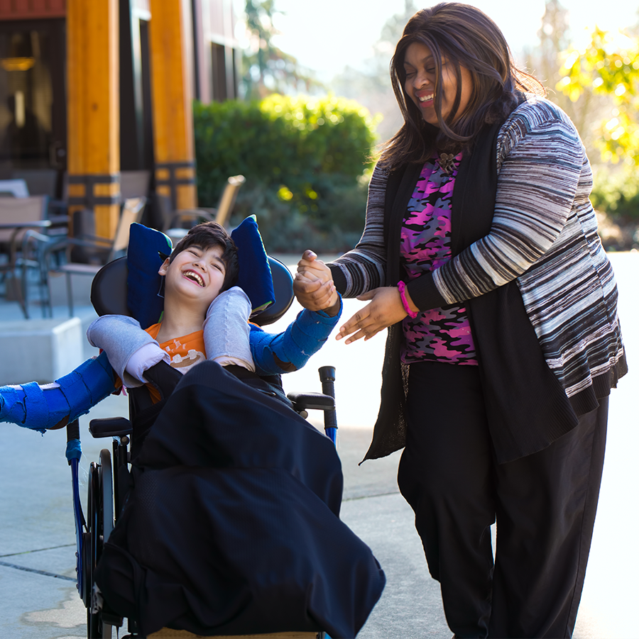 A happy Latinx girl in a wheelchair is holding hands with an African American women as they travel down a sidewalk