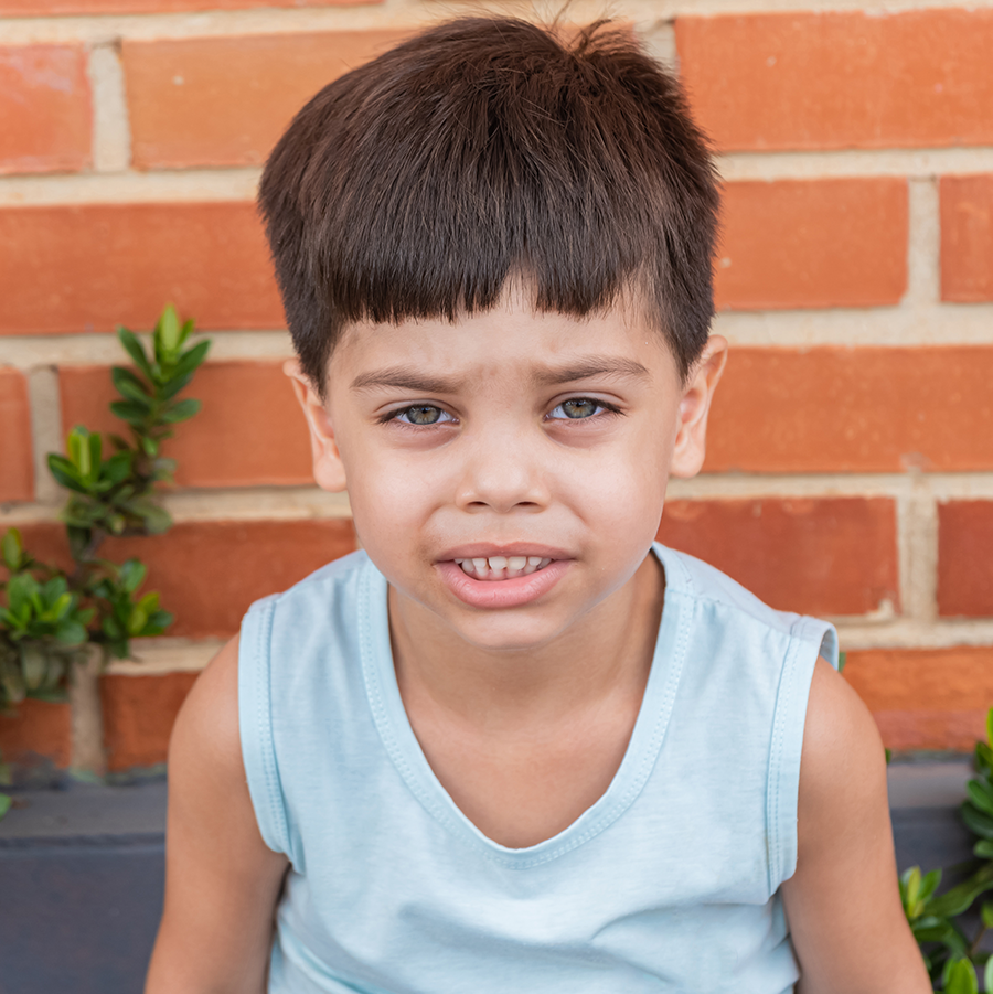 Young, Hispanic boy looking directly at the camera. He is wearing a blue shirt and standing in front of a brick wall.