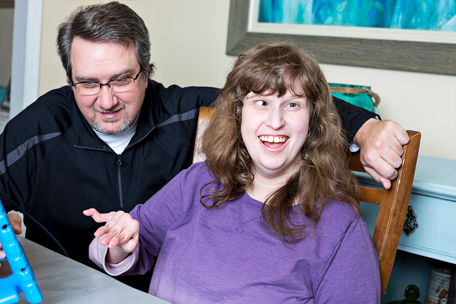 A man in a black jacket kneels next to a woman with disabilities sitting in a chair at a table