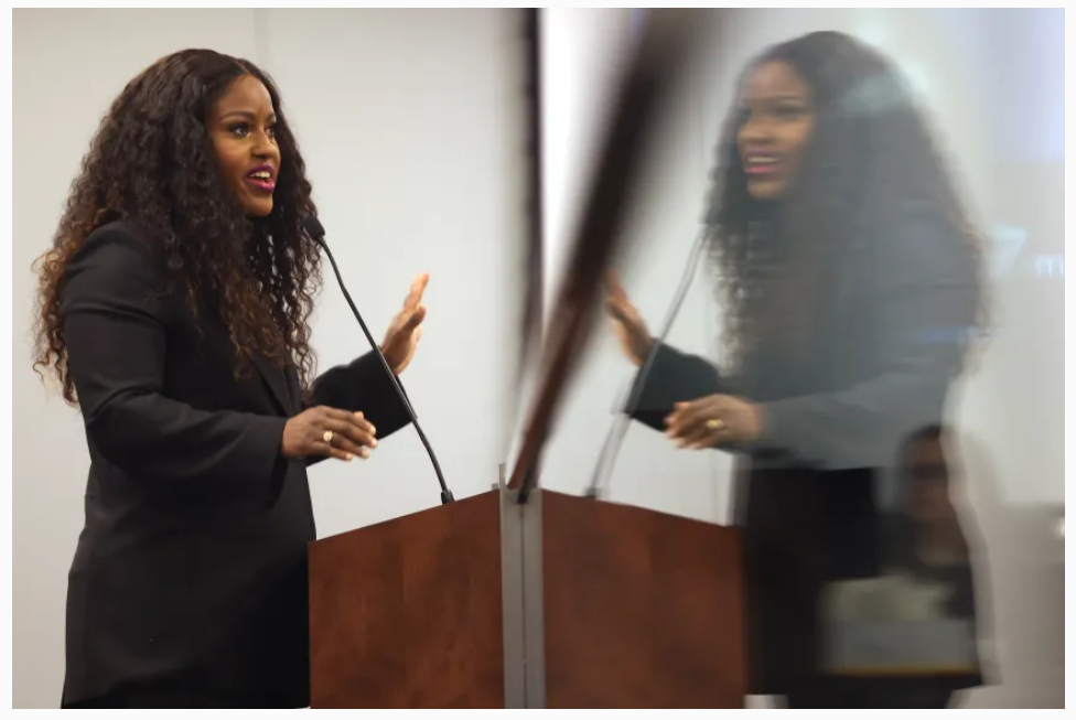 Photo from Chicago Tribune shows Stacey Davis Gates speaking at a podium with a mirror image reflected in plexiglass.