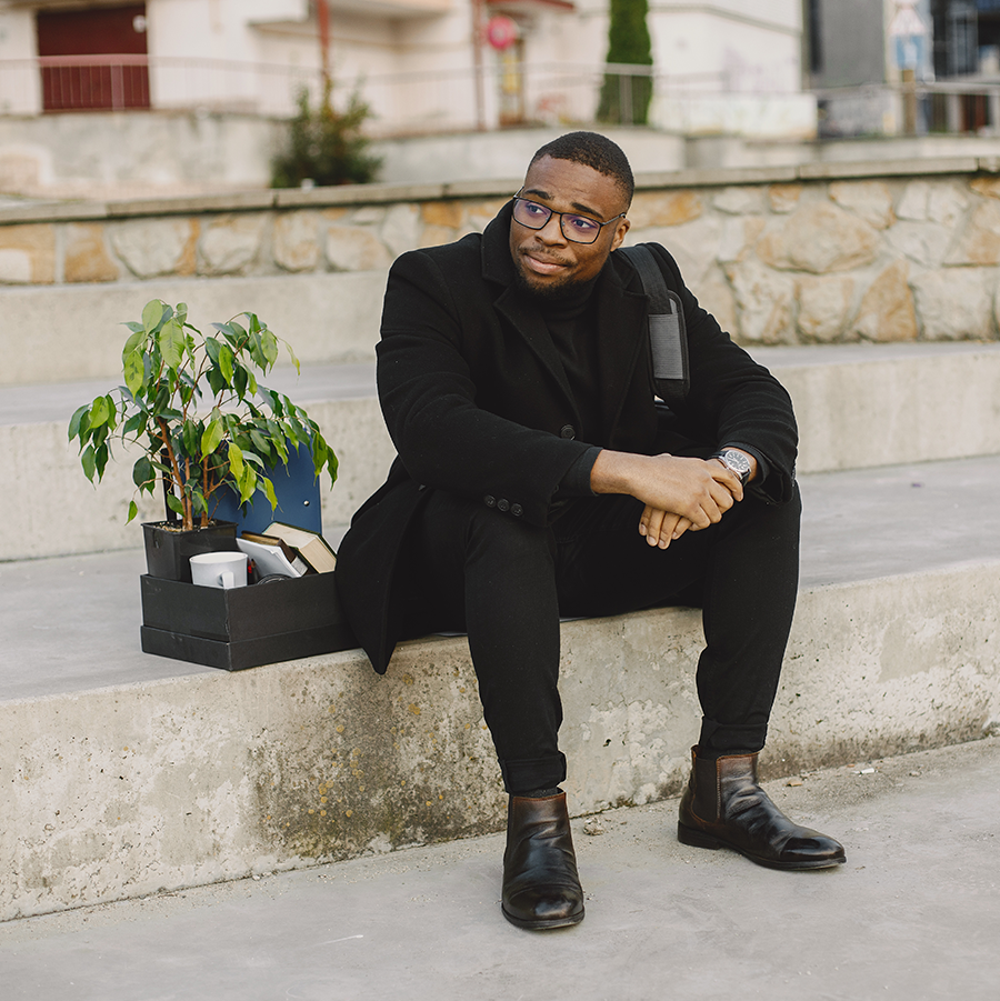 African American man in a suit sits on concrete steps with a box of work supplies next to him