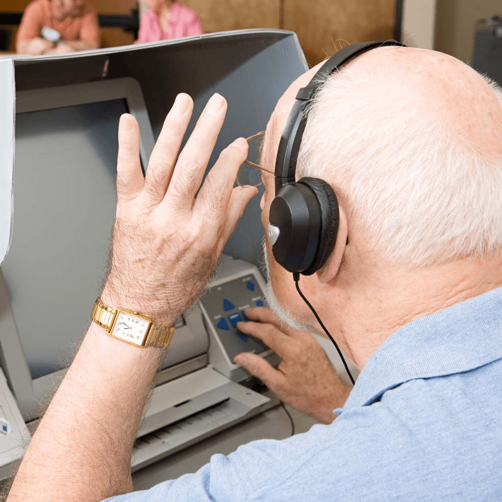 Elderly white man in a blue shirt looks closely at the screen of an accessible voting machine